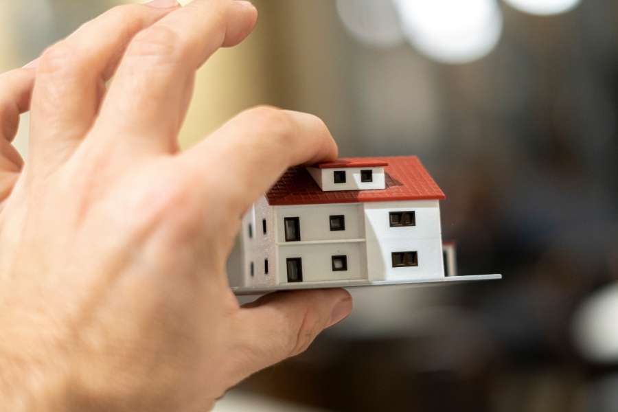 Une main tient délicatement une petite maquette de maison blanche au toit rouge pour illustrer le dispositif Pinel.